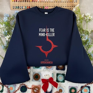 May include: Navy blue sweatshirt with the text "FEAR IS THE MIND-KILLER" in white. Below the text is a red graphic and the words "HOUSE HARKONNEN" in white. The sweatshirt has long sleeves.