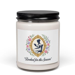 May include: A white candle jar with a black lid. The label features a gold-framed illustration of a person reading, surrounded by flowers. The text "Booked for the Season" is below the illustration. The candle is a home decor item.