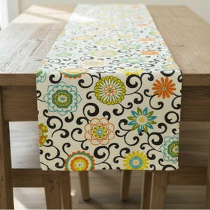 May include: A rectangular table runner featuring a cream-coloured background with a repeating pattern of black swirling vines and stylised flowers in shades of orange, lime green, and turquoise blue, arranged in a dense, symmetrical floral design.