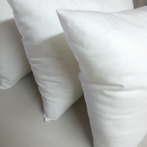 May include: A collection of white square pillows, made from a soft fabric, are arranged on a light surface. The pillows have a slightly textured appearance. These pillows are likely for home decor or bedding.