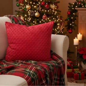 May include: A red textured cushion with a dotted pattern rests on a white sofa, covered with a red, green, and white plaid throw. A Christmas tree with decorations and lights is in the background, alongside lit candles and wrapped presents.