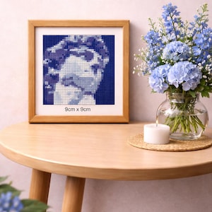 Peut inclure: Une œuvre d'art encadrée en point de croix représentant un buste classique en nuances de bleu, mesurant 9 cm x 9 cm. Elle est posée sur une table ronde en bois, avec un vase de fleurs bleues et une bougie blanche allumée.