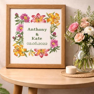 May include: A framed cross-stitch artwork featuring a floral wreath in shades of pink, yellow, and green. The text "Anthony & Kate" and the date "02.06.2022" are centered within the wreath. A vase of flowers and a candle are also visible.