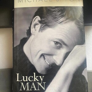 May include: A book titled "Lucky Man" by Michael J. Fox. The cover features a black and white portrait of a smiling man. The title is in large white letters, with "a memoir" below.