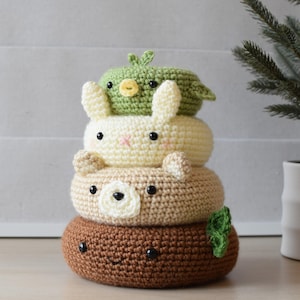 Puede incluir: Una pila de cuatro juguetes de animales de ganchillo. El juguete inferior es marrón con una cara sonriente y una hoja verde. Encima hay un oso marrón, luego un conejito crema y un pájaro verde. Cada uno tiene ojos negros.