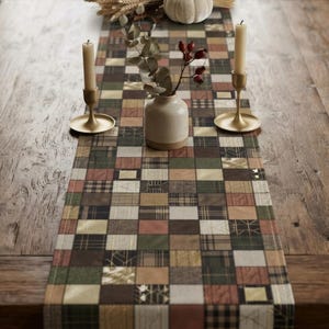 May include: A rectangular table runner featuring a patchwork design of various squares in shades of green, brown, cream, and muted red. The runner is decorated with two gold-toned candle holders with cream candles and a small white ceramic vase.
