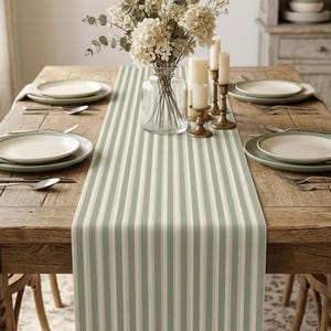 May include: A sage green and white striped table runner centered on a rustic wooden dining table. The table is set with four place settings, each featuring a white plate on a sage green charger, a silver fork, and a silver spoon.
