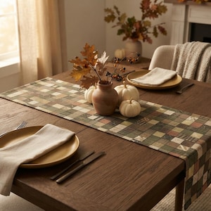 May include: A rectangular table runner featuring a patchwork pattern of squares in shades of olive green, brown, cream, and tan. The runner is placed on a dark wood dining table with two beige plates, white cloth napkins, and a ceramic vase with pumpkins.