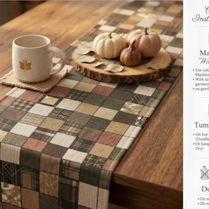 May include: A rectangular table runner featuring a patchwork pattern of squares in shades of brown, green, tan, and cream, with some squares displaying metallic gold geometric lines. The runner is displayed with a ceramic mug and decorative pumpkins.