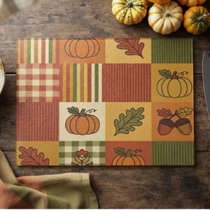 Puede incluir: Una tabla de cortar rectangular de vidrio con un diseño de mosaico de cuadrados con temática otoñal. El patrón incluye ilustraciones de calabazas naranjas, bellotas marrones y hojas de roble verdes, intercaladas con bloques a cuadros y rayas en colores de otoño.