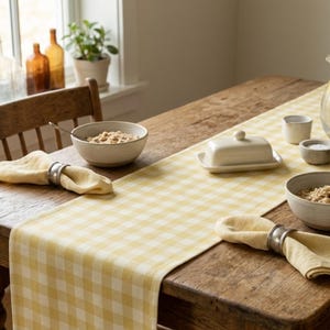 May include: A yellow and white gingham table runner laid across a rustic wooden table, featuring two light yellow cloth napkins in metal rings, two ceramic bowls with oatmeal, a covered butter dish, a small creamer, and a glass pitcher of water.