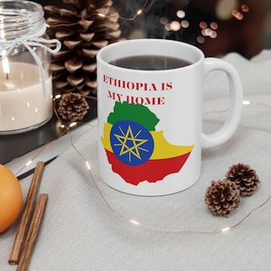 Pode incluir: Uma caneca de cerâmica branca com o texto ETHIOPIA IS MY HOME em letras maiúsculas vermelhas sobre um mapa da Etiópia com as cores da bandeira nacional verde, amarelo e vermelho, e um círculo azul central com uma estrela amarela.