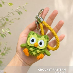 Può includere: Portachiavi a forma di gufo all'uncinetto nei toni del verde e dell'arancione, con cinturino intrecciato giallo e finiture argentate. Il gufo ha grandi occhi bianchi e un piccolo becco arancione. Il testo sull'immagine recita "CROCHET PATTERN" e "CREATED BY @EMBRILLY."