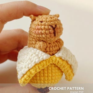 Può includere: Una figura di orso all'uncinetto che esce da una buccia di banana. L'orso è marrone chiaro con guance rosa e dettagli ricamati marroni. La buccia di banana è gialla e bianca. Il testo "CROCHET PATTERN" e "Digital PDF" sono visibili.