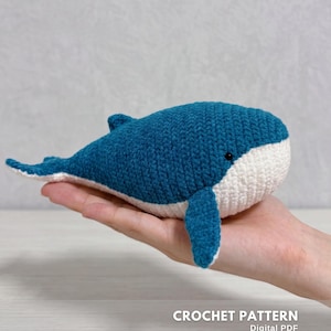Può includere: Un piccolo giocattolo a forma di balena realizzato all'uncinetto a mano, con il corpo superiore blu scuro e la pancia bianca. La balena ha un occhio nero visibile e una piccola pinna dorsale. Il testo CROCHET PATTERN Digital PDF CREATED BY @EMBRILLY è visibile.