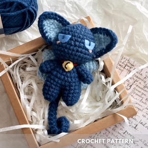 Può includere: Una bambola a forma di gatto all'uncinetto blu scuro fatta a mano, con dettagli azzurri, grandi orecchie e un collare con campanello. La bambola è presentata in una piccola scatola di legno con imbottitura di carta, accanto a un gomitolo di lana blu. Il testo "CROCHET PATTERN" è presente.