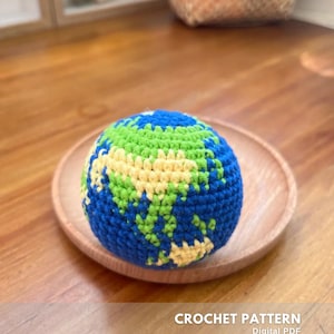 Peut inclure: Un globe terrestre crocheté, avec des océans bleus, des continents verts et jaunes, et des détails blancs. La sphère faite à la main est posée sur une assiette en bois. Le texte en bas indique "CROCHET PATTERN Digital PDF CREATED BY @EMBRILLY".