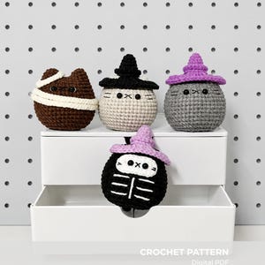 Puede incluir: Cuatro peluches de ganchillo redondos con caras de gato. Uno es marrón con vendas blancas, uno beige con sombrero de bruja negro, uno gris con sombrero de bruja morado y uno negro con diseño de caja torácica de esqueleto y sombrero morado.
