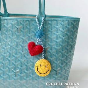 Può includere: Una borsa tote turchese con un motivo bianco e blu, con un ciondolo all'uncinetto fatto a mano. Il ciondolo include un cerchio blu, un cuore rosso e una faccina sorridente gialla. Il testo "CROCHET PATTERN" è visibile.