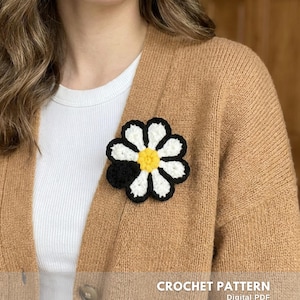 Può includere: Spilla a forma di margherita all'uncinetto con petali bianchi, centro giallo e bordo nero, appuntata su un cardigan beige. Il testo "CROCHET PATTERN Digital PDF CREATED BY @EMBRILLY" è visibile in basso.