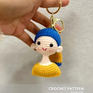 May include: Crocheted keychain featuring a doll with a blue hat, yellow top, and pearl earrings. The keychain has a gold-colored ring and clasp. The text "CROCHET PATTERN" is visible at the bottom of the image.