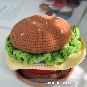 May include: A crocheted burger with a brown bun sprinkled with white stitches, green lettuce, yellow cheese, and a brown patty. The text "CROCHET PATTERN" is visible at the bottom of the image.