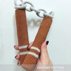 Può includere: Una coppia di nunchaku all'uncinetto marroni collegati da una catena all'uncinetto grigio chiaro. Ogni manico presenta due strisce orizzontali grigio chiaro vicino alla base. Il testo CROCHET PATTERN Digital PDF CREATED BY @EMBRILLY è visibile.