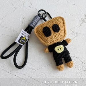 Puede incluir: Un llavero de crochet hecho a mano con diseño de robot. El robot tiene una cabeza beige con ojos negros, un cuerpo negro y pies beige. Está unido a un cordón negro con un acento gris y un cierre de metal. El texto "CROCHET PATTERN" es visible.