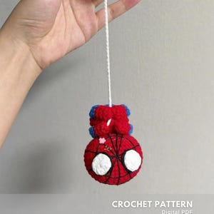 May include: A handmade crochet Spider-Man ornament. The ornament is red with a black web design and white eyes. It has blue accents and is suspended by a white string. The text "CROCHET PATTERN" is visible.
