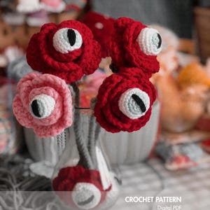 May include: A crochet pattern digital PDF created by @embrilly featuring four crocheted flowers in red and pink, each with a central white and light blue eyeball with a black vertical pupil, displayed in a clear glass vase.