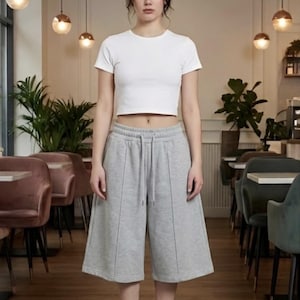 Könnte beinhalten: Ein weißes kurzärmeliges bauchfreies Oberteil kombiniert mit grauen knielangen Culotte-Shorts mit Kordelzug und vertikalen Nahtdetails, getragen mit weißen Söckchen und weißen Turnschuhen.