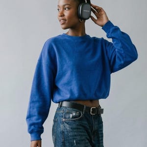 May include: A vibrant royal blue long-sleeve cropped sweatshirt with a crew neckline, paired with dark blue denim jeans featuring a black leather belt with a silver-toned metal buckle, and dark gray over-ear wireless headphones.