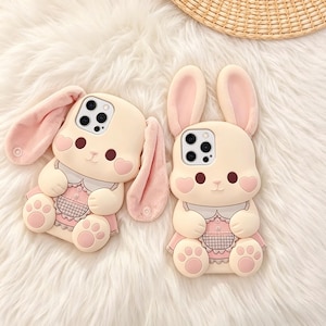 May include: Two phone cases shaped like adorable bunnies. The cases are primarily beige with pink accents, including the inner ears, heart-shaped cheeks, and the dress. The bunny's face features dark brown eyes and a small nose. The cases are on a white, fluffy surface.