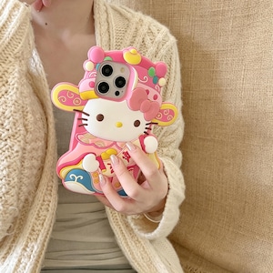 May include: Pink phone case featuring a Hello Kitty design with a festive, ornate outfit. The case has a 3D design with yellow, blue, and white accents. The phone case is designed to fit a smartphone.