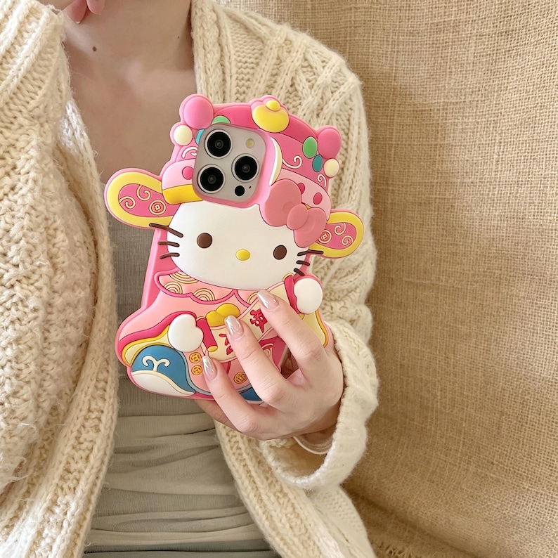 May include: Pink phone case featuring a Hello Kitty design with a festive, ornate outfit. The case has a 3D design with yellow, blue, and white accents. The phone case is designed to fit a smartphone.