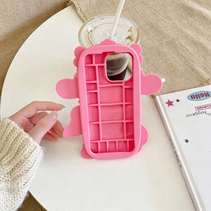 May include: A bright pink phone case with a cartoon character design. The case has a grid-like interior and features rounded edges and protruding shapes. A clear plastic cup with a straw and a notebook with the word "Hello" are also visible.