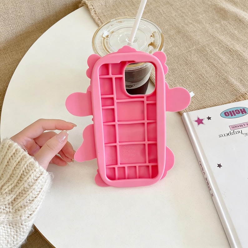 May include: A bright pink phone case with a cartoon character design. The case has a grid-like interior and features rounded edges and protruding shapes. A clear plastic cup with a straw and a notebook with the word "Hello" are also visible.