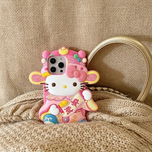 May include: Pink phone case featuring a cartoon character with a bow, wearing a traditional outfit. The case has a white face, black whiskers, and a yellow nose. The case has a gold handle and is resting on a beige knitted surface.