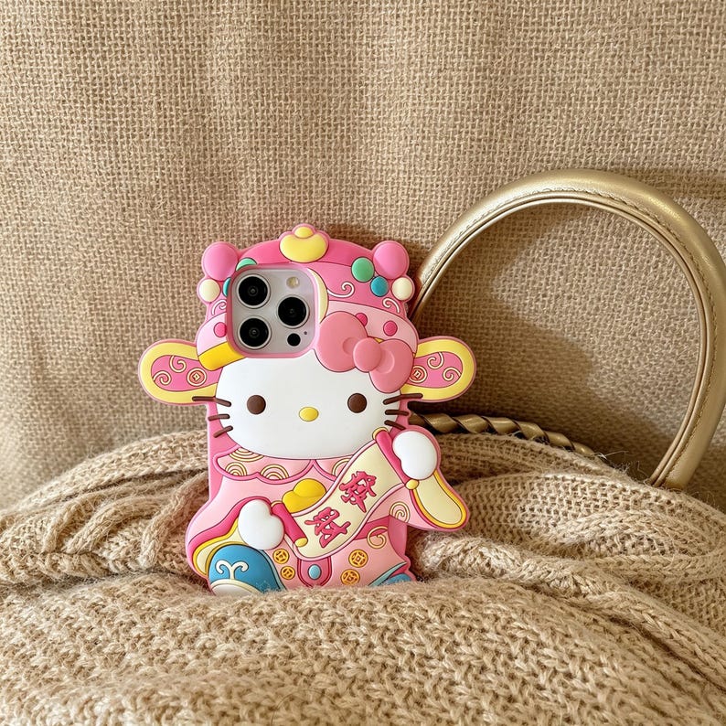 May include: Pink phone case featuring a cartoon character with a bow, wearing a traditional outfit. The case has a white face, black whiskers, and a yellow nose. The case has a gold handle and is resting on a beige knitted surface.
