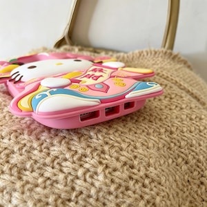 May include: A pink phone case featuring a cartoon character design. The case has a white face, pink body, and colorful accents. The design includes text and decorative elements. The case is made of a soft, flexible material.
