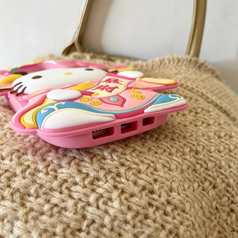 May include: A pink phone case featuring a cartoon character design. The case has a white face, pink body, and colorful accents. The design includes text and decorative elements. The case is made of a soft, flexible material.