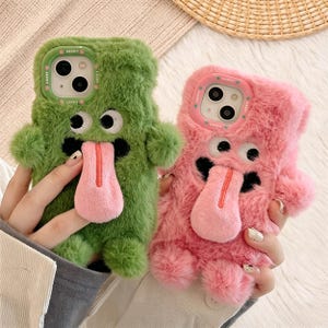 Op de afbeelding: Twee pluizige telefoonhoesjes, één groene en één roze, elk ontworpen om op een cartoonmonster te lijken. De hoesjes hebben grote wiebelogen, zwarte accenten en een roze tong. Op het groene hoesje staat het woord "Lucky".