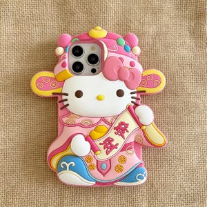 May include: Pink phone case featuring Hello Kitty in a traditional Chinese outfit. The case has a bow, a scroll with Chinese characters, and yellow and blue accents. The case is designed to fit a smartphone.