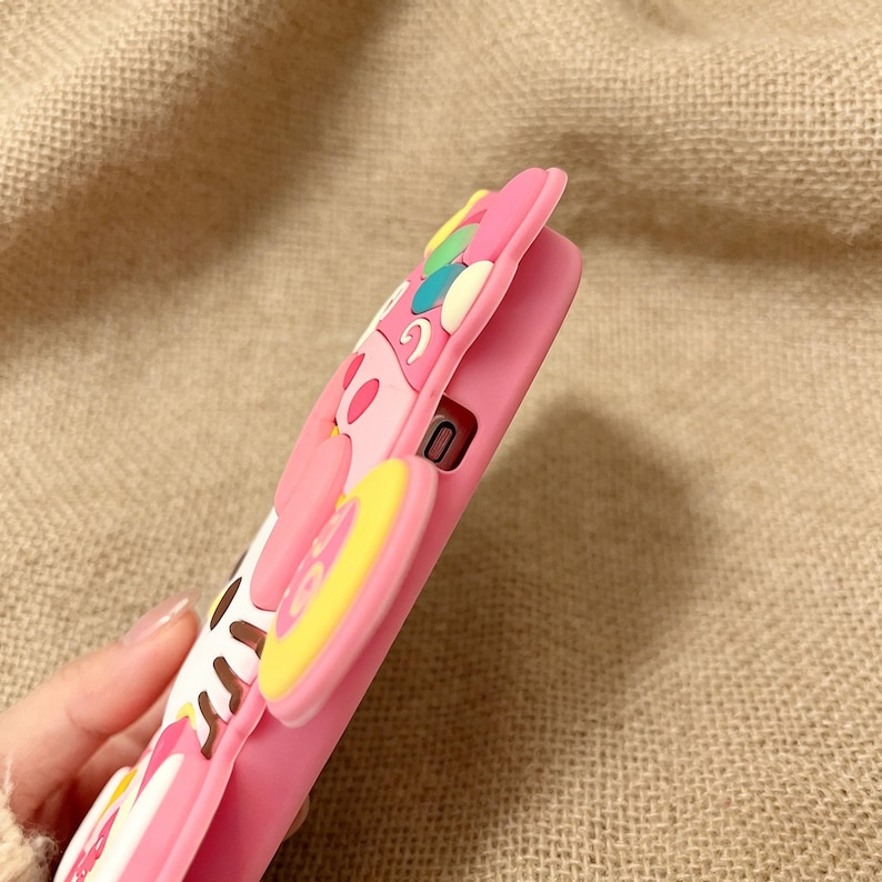 May include: Pink phone case with a colorful cartoon design. The case features a raised, three-dimensional design with a pink background and accents of yellow, white, and brown. The case is made of a soft, flexible material.