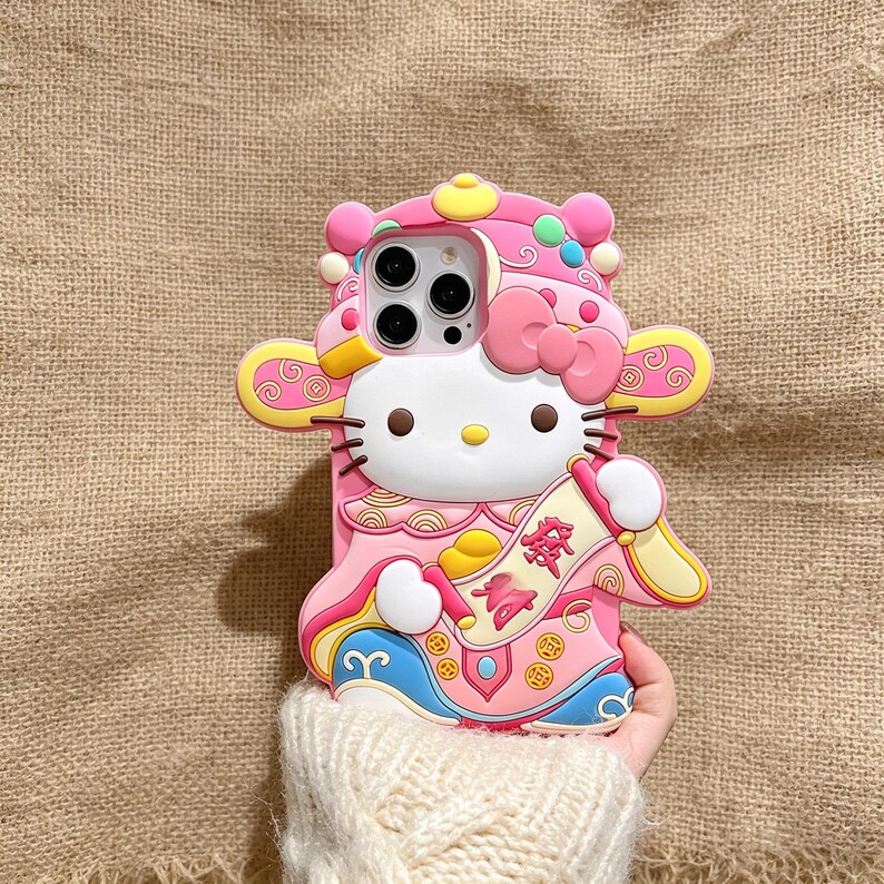 May include: A pink and white phone case featuring Hello Kitty in a traditional Chinese outfit. The case has a bow, decorative accents, and a scroll with Chinese characters. The case is made of a soft material.