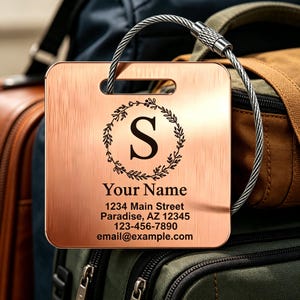 May include: A copper luggage tag with a brushed metal effect. The tag displays the letter "S" within a laurel wreath, with the words "Your Name" and contact details below. A silver cable loop is attached.