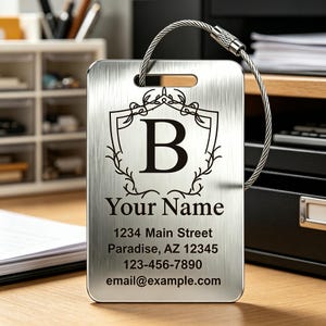 May include: A rectangular brushed silver metal luggage tag featuring a black printed crest with the letter B, the text Your Name, and placeholder contact details including 1234 Main Street, Paradise, AZ 12345, 123-456-7890, and email@example.com, with a steel cable loop.