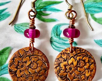 May include: A pair of copper earrings with a floral design and pink glass beads. The earrings are hanging from silver hooks.