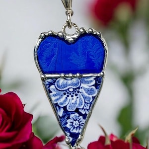 May include: A silver-toned necklace with a pendant featuring a blue heart-shaped top and a triangular bottom with a white and blue floral pattern. The pendant is surrounded by a silver frame.