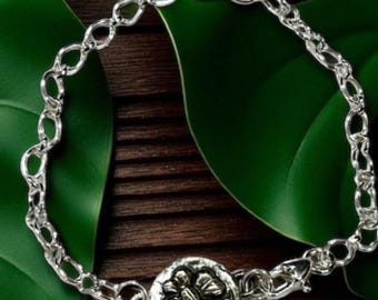 Può includere: Un braccialetto argentato con una catena a maglie ovali e un elemento centrale floreale circolare. Il braccialetto è presentato su una superficie di legno scuro con foglie verdi. Ha una chiusura a moschettone.